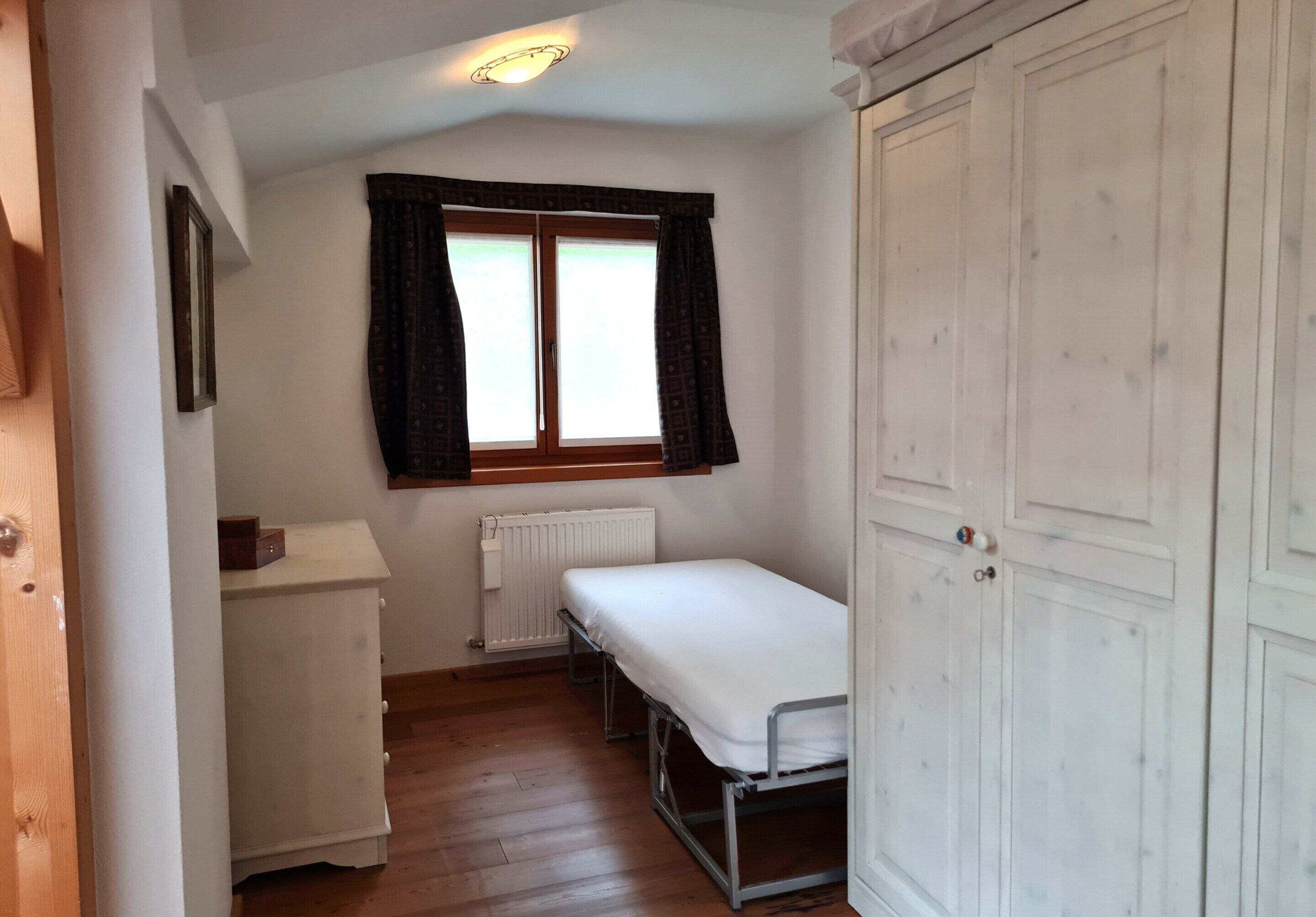 A small, single-room hotel room with a wooden floor, a white bed, a bedside table, a radiator, and a large wardrobe.