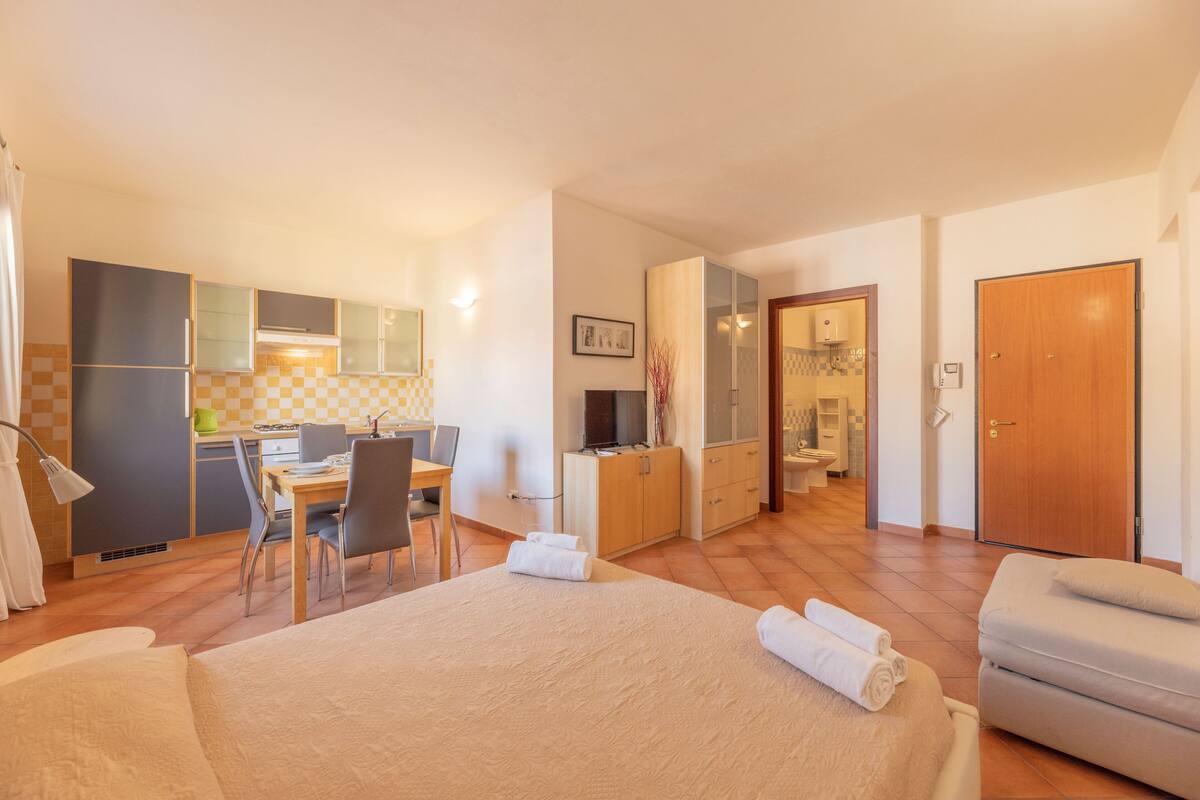 A hotel room with a bed, a desk, a chair, a kitchenette, and a bathroom.