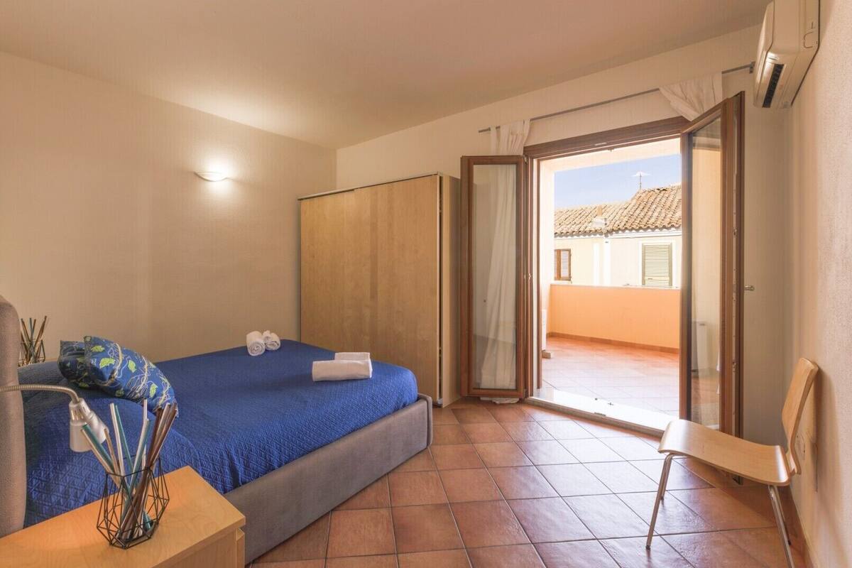 A bedroom with a bed, a chair, a bedside table, a wardrobe, and a sliding glass door leading to a balcony.