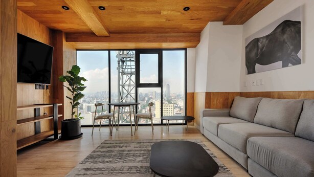 Ein modernes Wohnzimmer mit grauem Sofa, Pouf in Schwarz, einem Holz-TV-Schrank und einem großen Fenster mit Blick auf die Stadt.