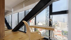 Ein modernes Zimmer mit schrägem Dach, einem Holztisch, einem Korbstuhl und großen Fenstern mit Blick auf die Stadt.