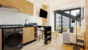 Eine moderne Küche mit integriertem Waschtrockner, eine Essecke mit einem kleinen Tisch und Stühlen sowie ein Sofa in einem Wohnbereich mit einem großen Fenster.