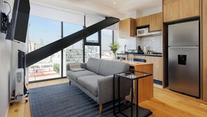 Ein modernes Hotelzimmer mit einer Sitzecke, einem kleinen Tisch, einer Küchenzeile mit Kühlschrank und einem großen Fenster mit Blick auf die Stadt.