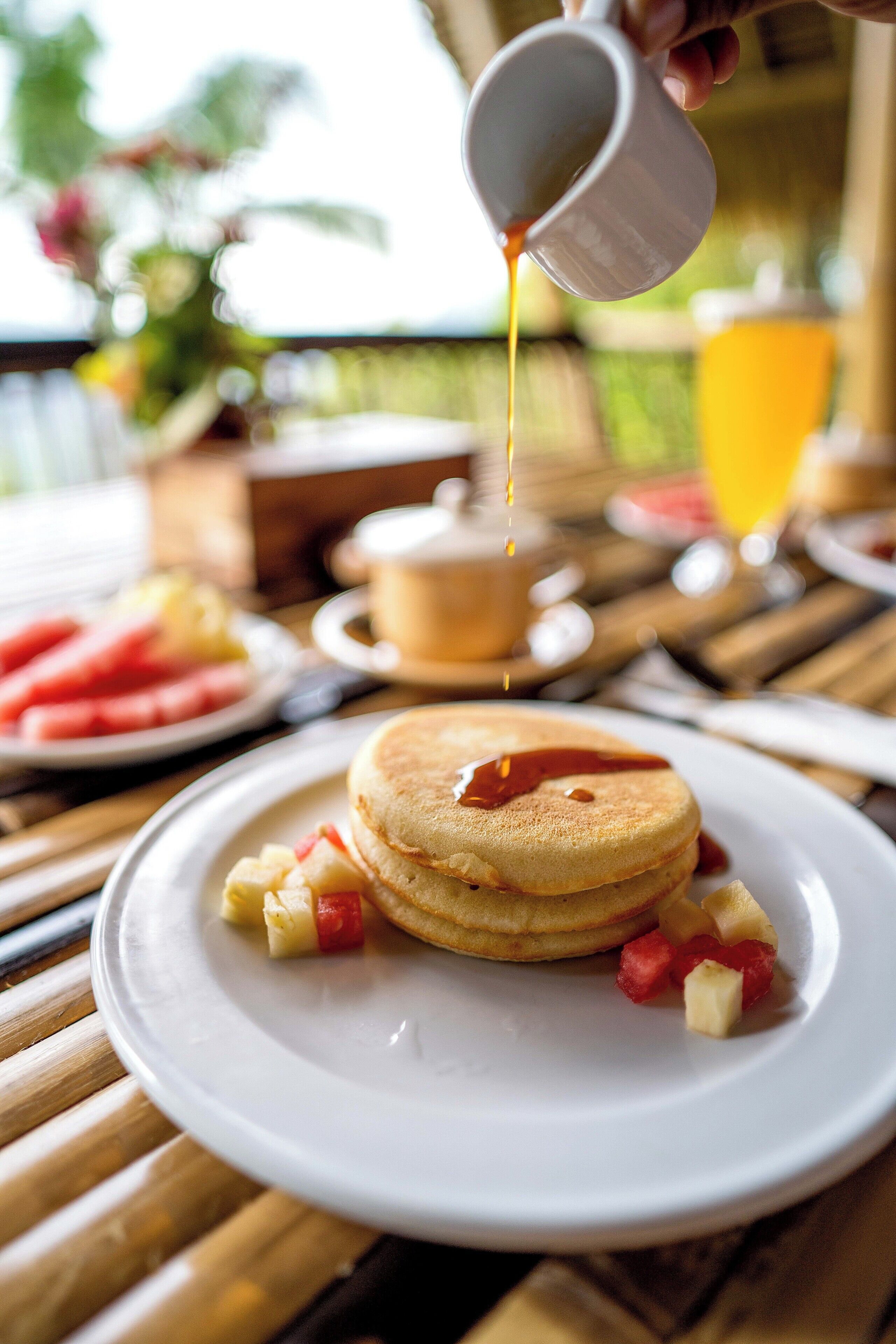 A breakfast setting with pancakes on a plate, syrup being poured, and a fruit plate in the background.