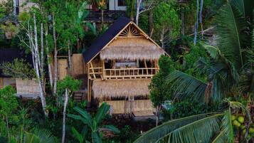 A thatched-roof structure surrounded by lush greenery, with a wooden balcony and a staircase leading to an upper level.