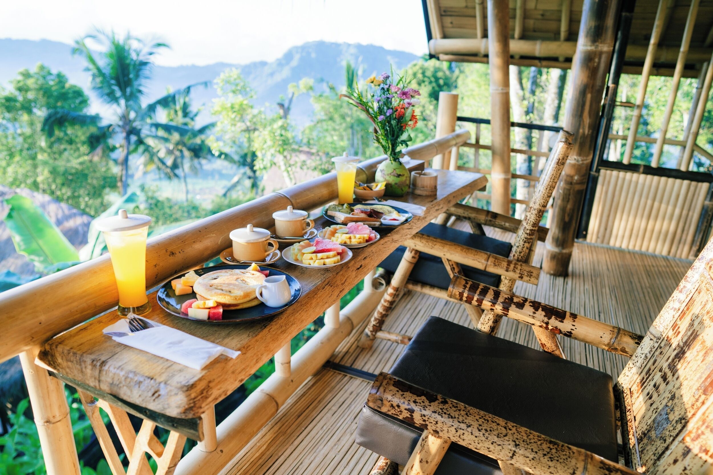 A wooden table with breakfast items and a glass of orange juice, set on a balcony with bamboo railings and a view of greenery and mountains.