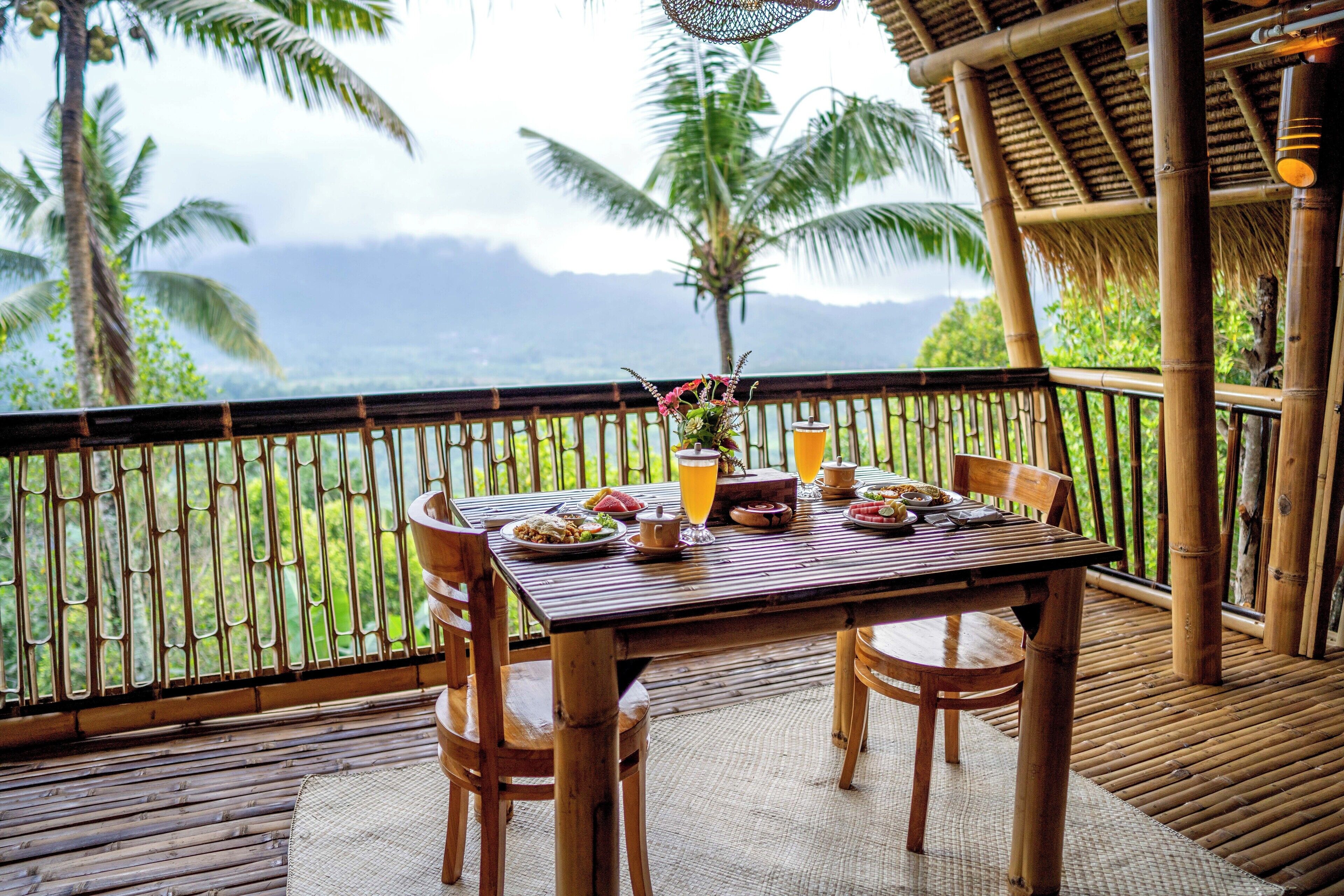 A wooden table set with plates, glasses, and a vase of flowers on a balcony with bamboo railings and a view of palm trees and mountains.