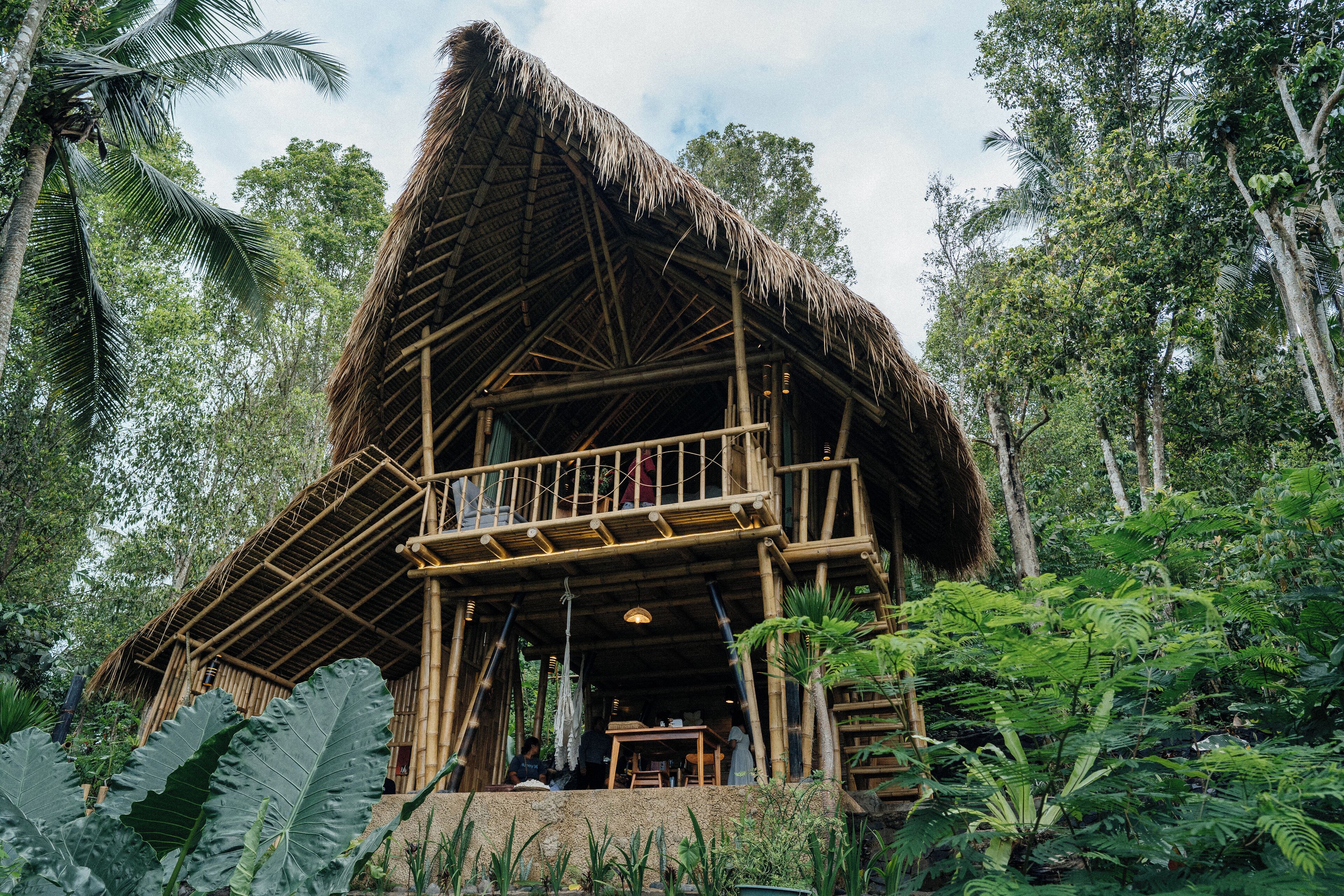 A bamboo structure with a thatched roof, a balcony, and surrounded by lush greenery.