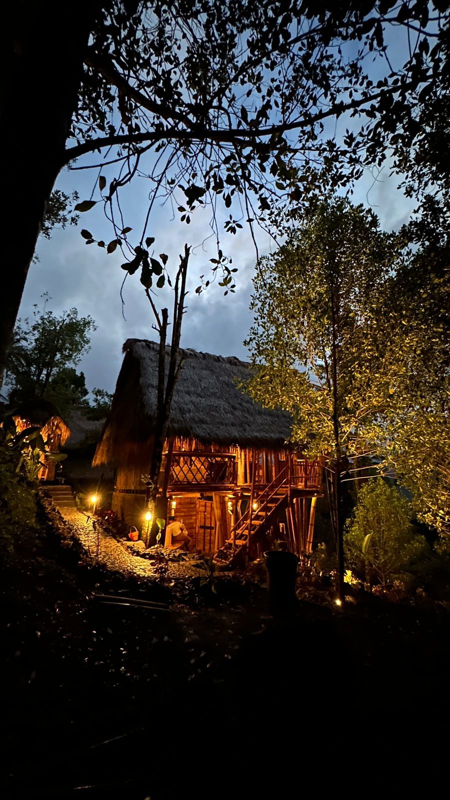 A rustic thatched-roof hut surrounded by dense foliage, with a wooden staircase leading up to it.