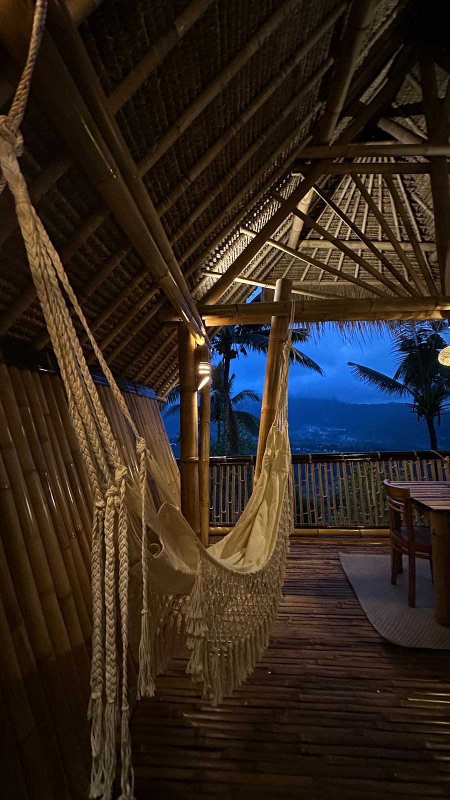 A hammock under a thatched roof structure with a wooden deck and a view of palm trees and mountains.