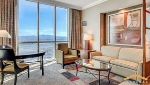 A hotel room with a large window offering a view of the city and mountains, a sofa, a chair, a small table, and a lamp.