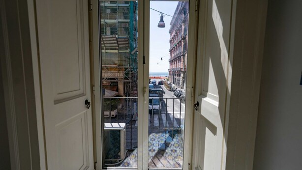 A view through a glass door of a building with a tiled floor, a metal railing, and a street with parked cars and buildings in the background. - Arcoleo Prestige 2 By Dimorra (NAPOLI)