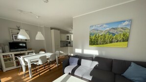 A modern living room with a grey sofa, a white coffee table, and a dining area with a white table and chairs. A kitchen with white cabinets and a microwave is visible in the background. - Exclusive Apartment With Scenic View (Bad Kleinkirchheim)