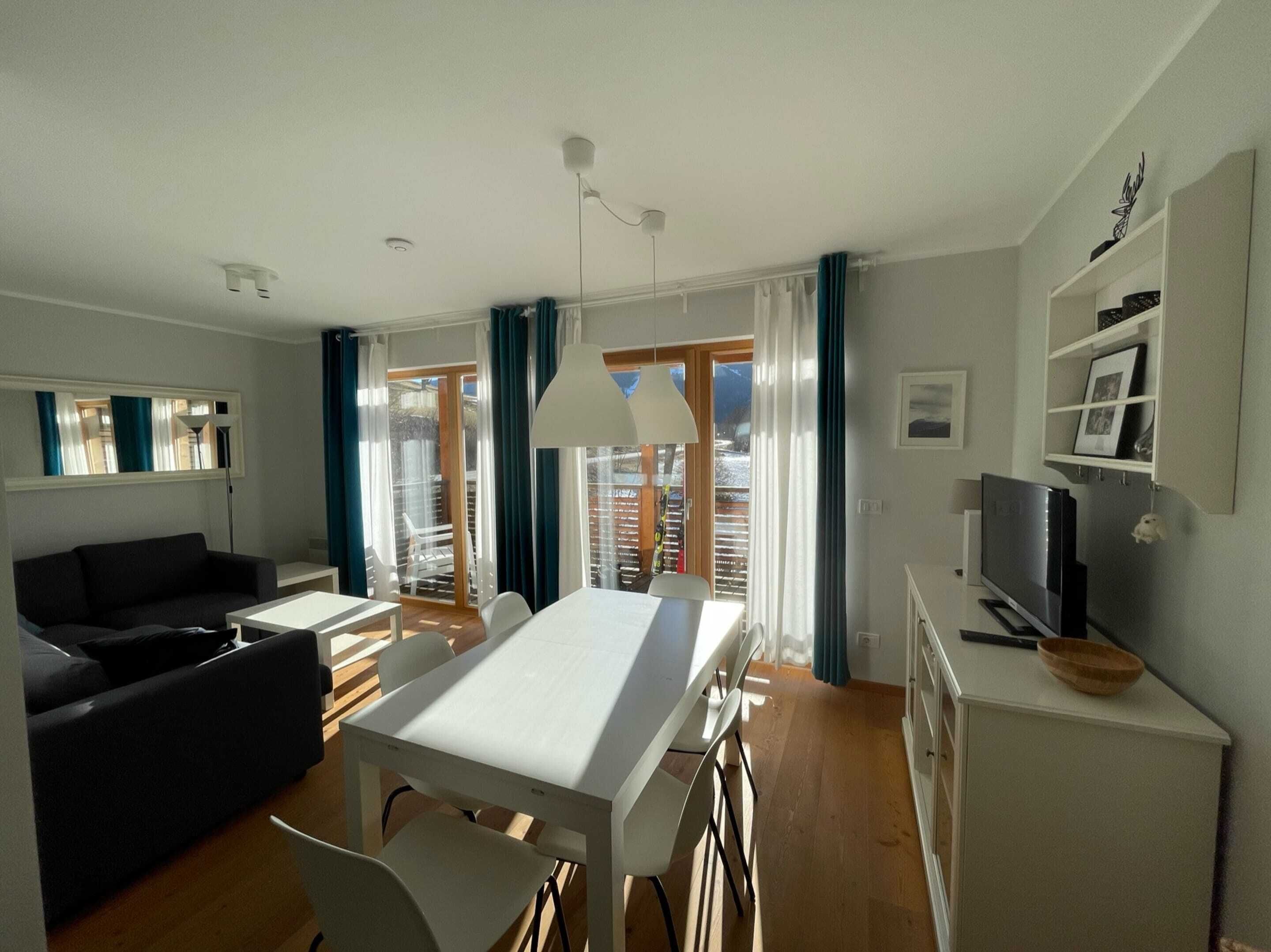 Una sala de estar moderna con comedor, sofá, televisor y un balcón con cortinas.