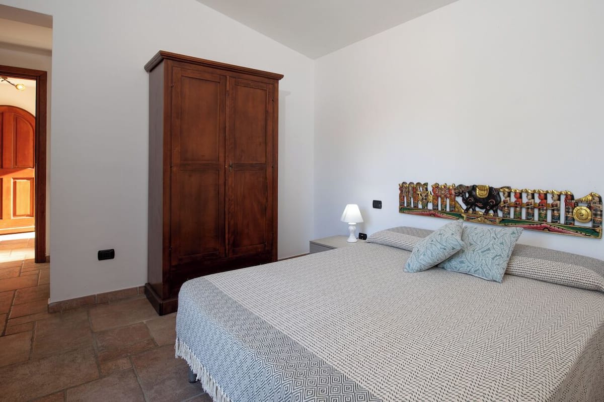 A bedroom with a large bed, a wooden wardrobe, a wall-mounted artwork, and a bedside lamp.