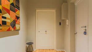 Un couloir avec un plancher en bois, une porte beige, un petit tabouret noir et un tableau abstrait aux couleurs vives accroché au mur.