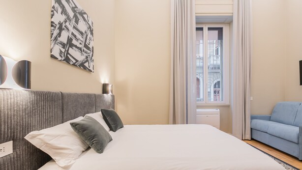 Une chambre d’hôtel bien rangée, avec un lit, un fauteuil bleu, un grand tableau abstrait et une fenêtre avec des rideaux.