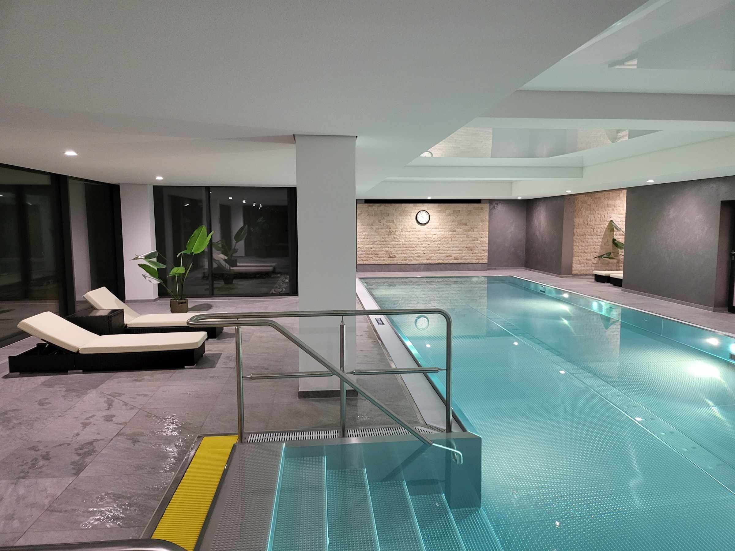 Ein moderner, überdachter Swimmingpool mit Glastrennwand, Liegestühlen und einer Uhr an der Wand.