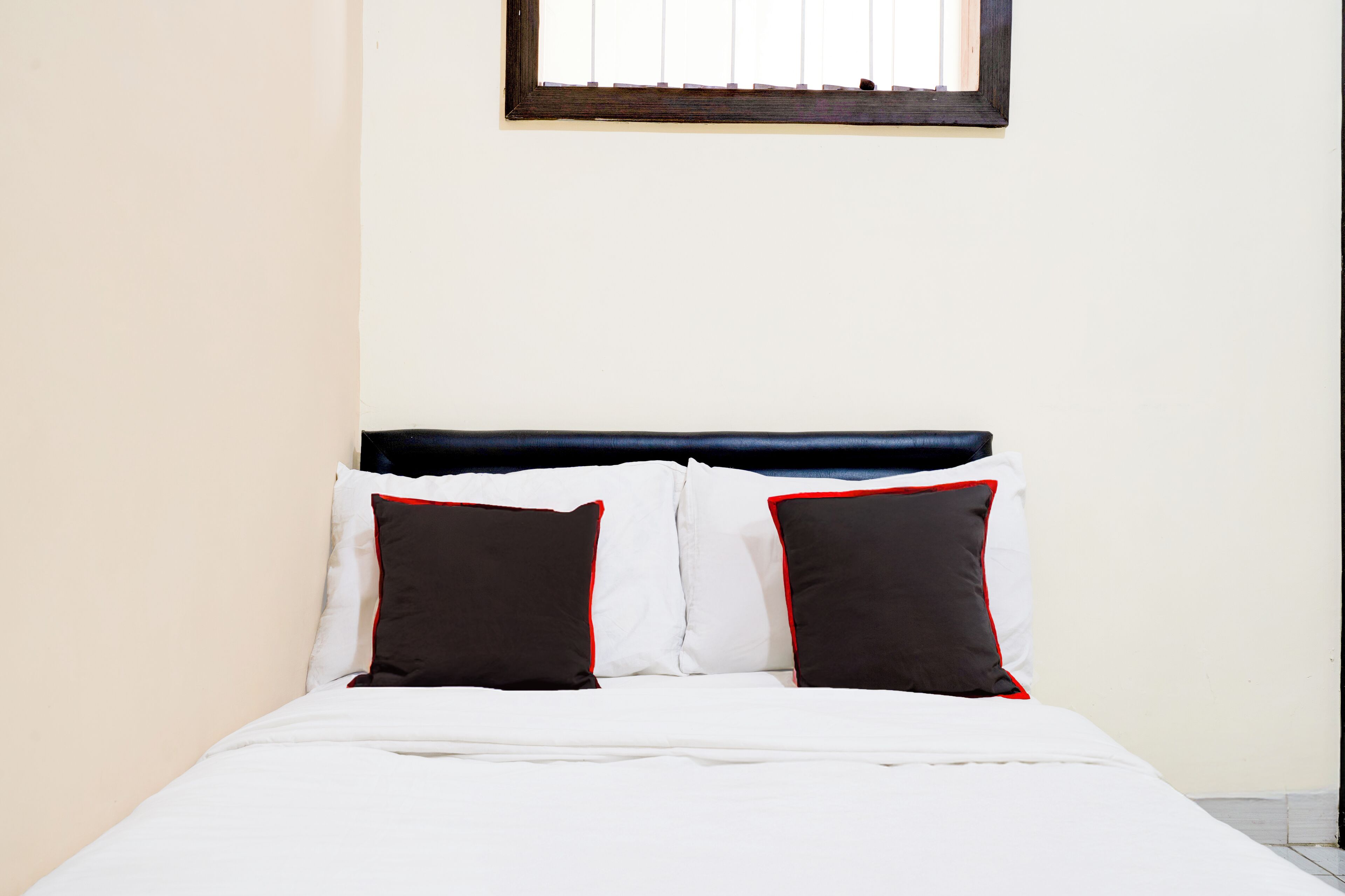 A bed with white bedding and two black pillows with red trim, a dark headboard, and a window with blinds.