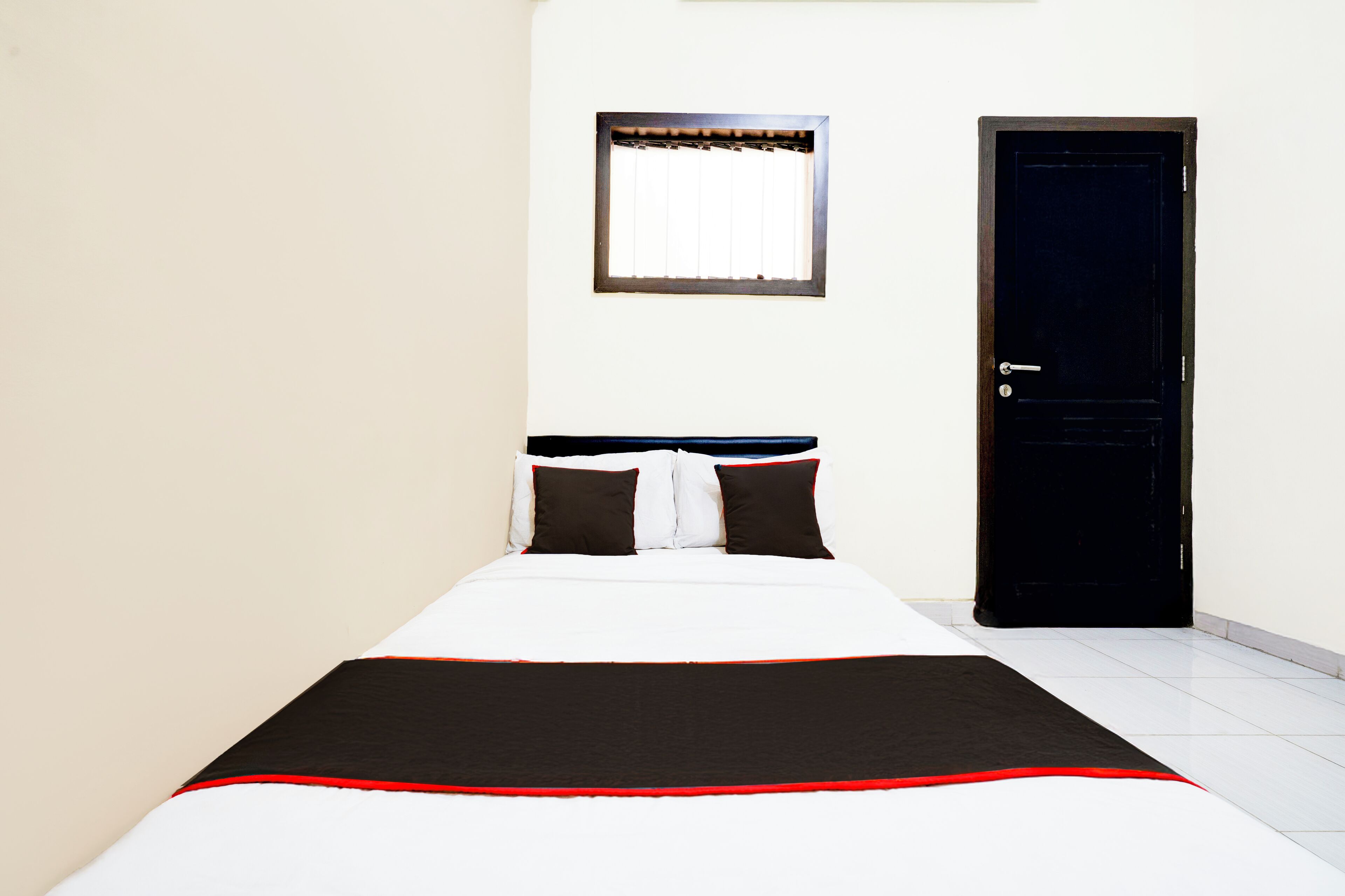 Un lit bien fait, recouvert d’une couvre-lit noire et blanche, une fenêtre avec des stores et une porte fermée.