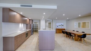 A modern kitchen with a central island, a dining area with a long table, and a large window with a view. - Casa Di Marco Polo - 3 Bedroom Apartment (Mandurah)