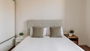 Ein ordentlich bezogenes Bett mit grauem Polsterkopfteil, zwei Nachttische mit Uhren und Pflanzen, sowie eine kleine Kommode unter dem Kopfteil.