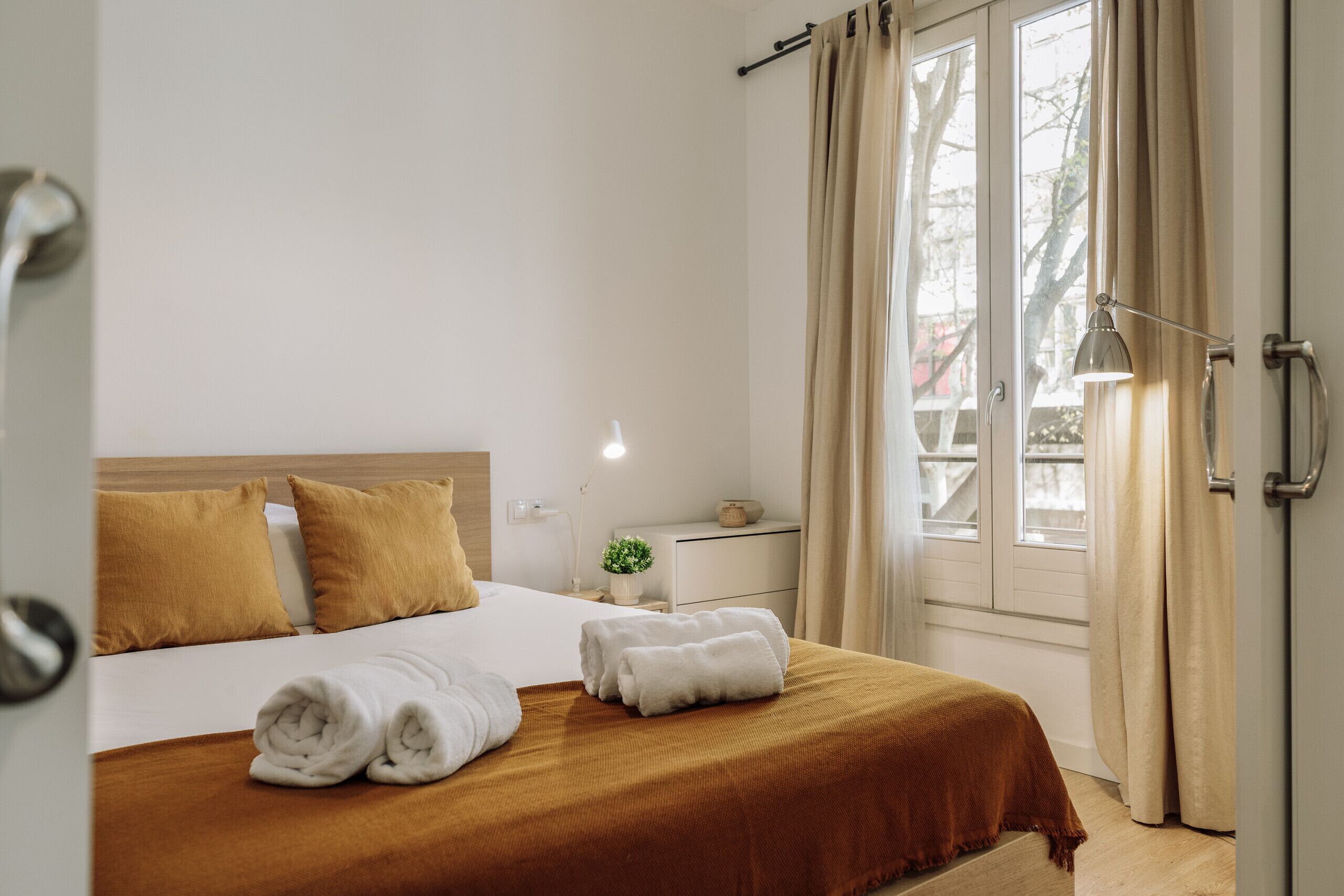 A hotel room with a bed, a bedside table, a lamp, and a window with curtains.
