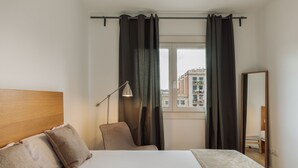 Ein Hotelzimmer mit Bett, Sessel, Schreibtisch, Lampe und einem Fenster mit Vorhängen.