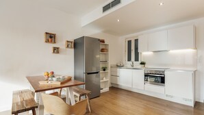 Eine moderne Küche mit weißen Schränken, einem Edelstahlkühlschrank und einem Backofen. Ein Holztisch mit Stühlen und ein kleiner Topfpflanze an der Wand sind ebenfalls vorhanden.