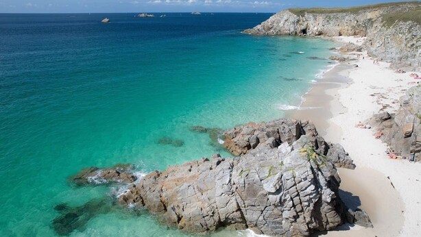 A coastal beach with clear turquoise waters, rocky outcrops, and a sandy shoreline.