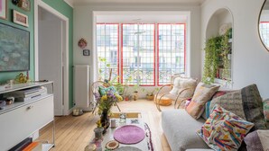 A cozy living room with a sofa, a coffee table, and a bookshelf. There is a large window with red frames and a view of the outside. - Behind the Stained Glass, by Veeve (Paris)