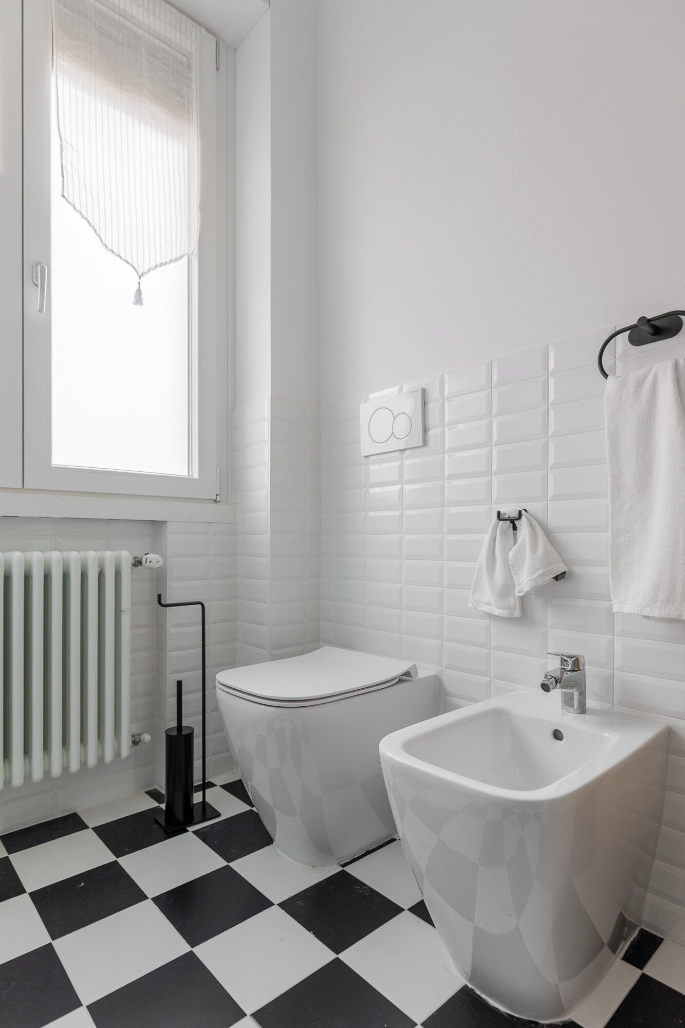 A bathroom with a black and white checkered floor, a toilet, a bidet, a sink, and a window with a white curtain.