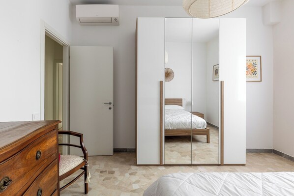 A bedroom with a wooden dresser, a wardrobe, a bed with a white duvet, and a ceiling fan.