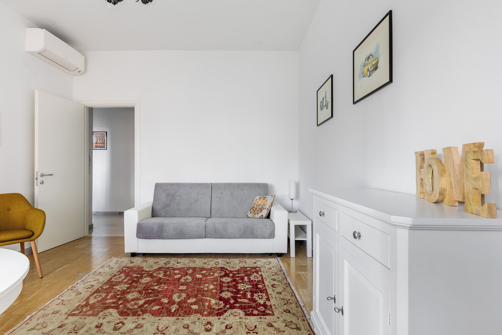 En moderne stue med en grå sofa, et rødt og beige tæppe med mønster, et hvidt skab og en gul stol.