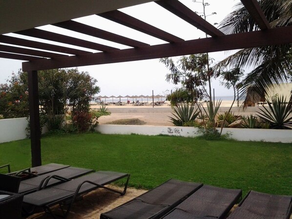 A covered patio with lounge chairs, a view of the beach, and a white wall with plants.