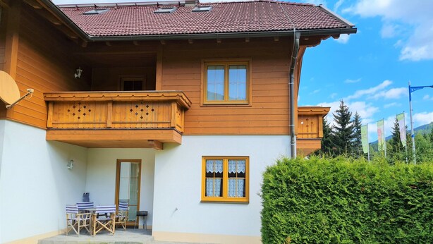 A two-story wooden house with a balcony, a small patio with a table and chairs, and a well-maintained hedge. - Bright Apartment Near Thermal Springs (Bad Kleinkirchheim)
