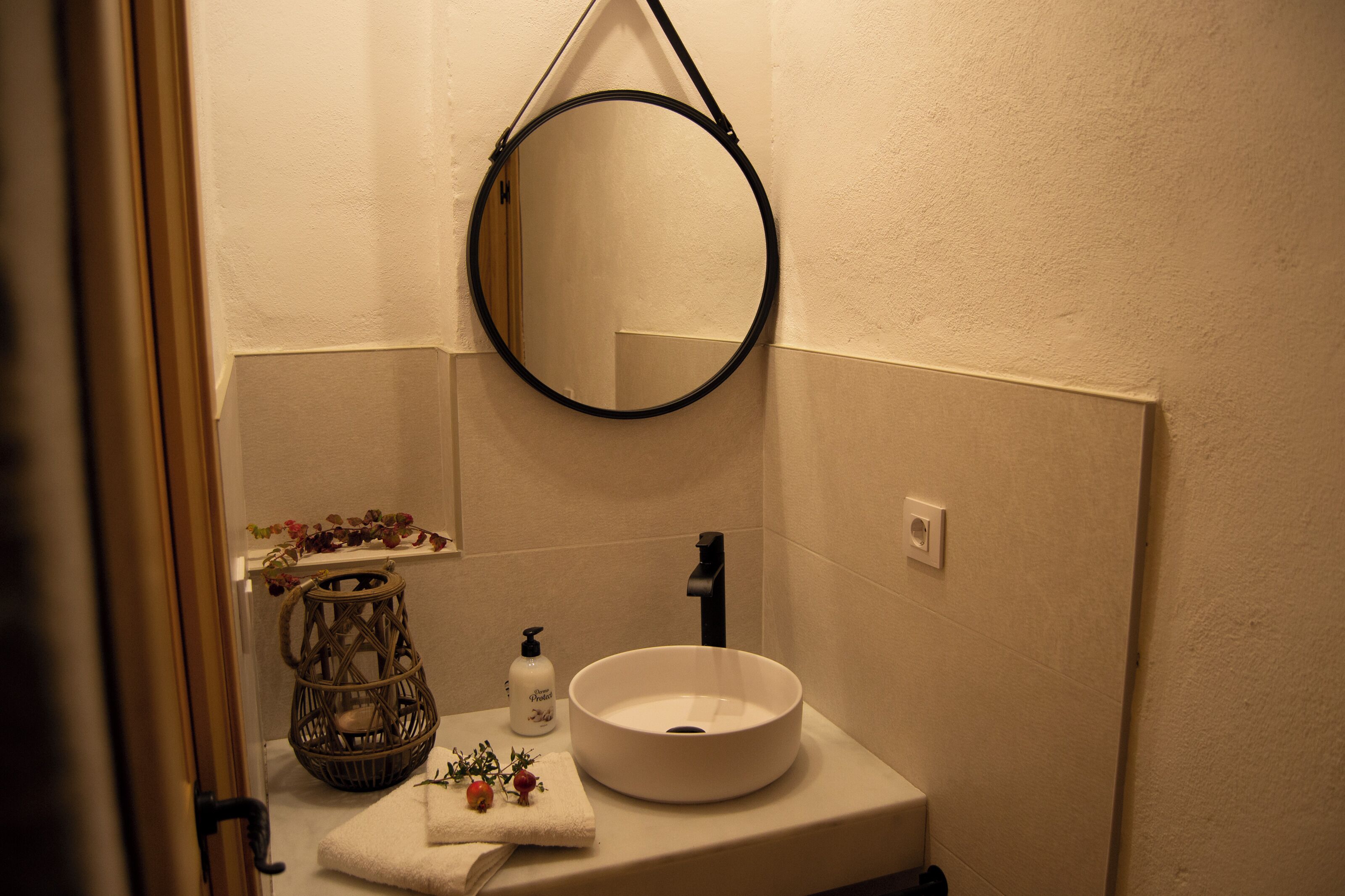 Une salle de bain avec un miroir rond, un lavabo blanc, un robinet noir, un panier en osier et un flacon de savon.