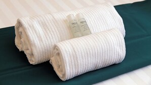 Zwei gerollte weiße Handtücher und zwei Flaschen Dermocare Hotel Care Produkte auf einem Bett mit grüner Bettdecke.