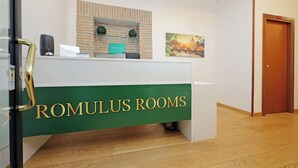 Ein Empfangsschalter mit der Beschriftung 'ROMULUS ROOMS', eine Wand mit Ziegelverkleidung, ein gerahmtes Bild und eine Holztür.