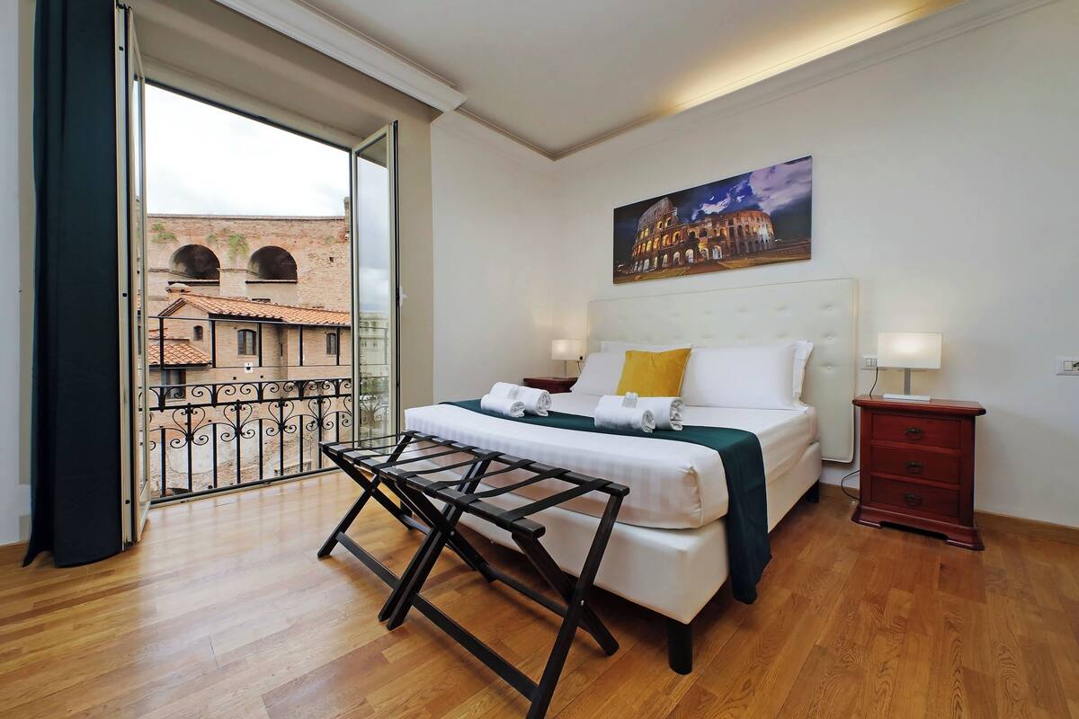 A hotel room with a bed, a nightstand, a lamp, a balcony with a view of a historic building.