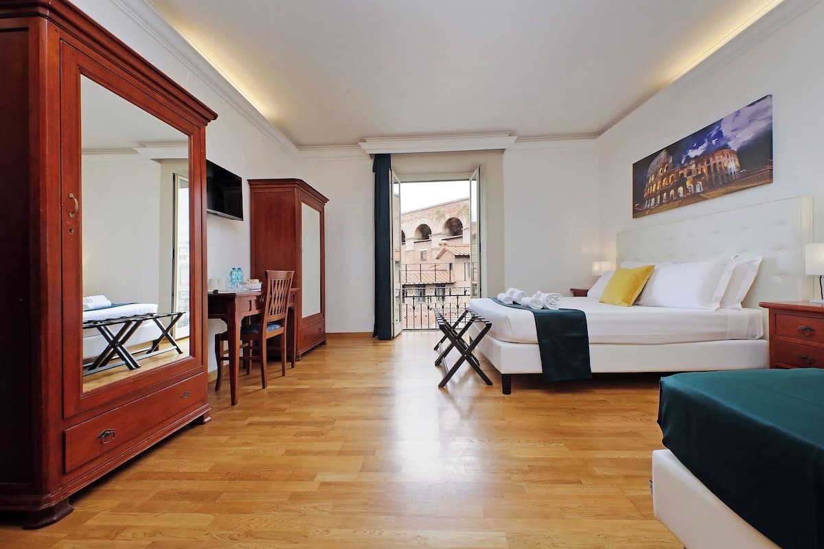 A hotel room with a wooden floor, a bed with white bedding, a desk with a chair, a large mirror, and a painting of a building on the wall.