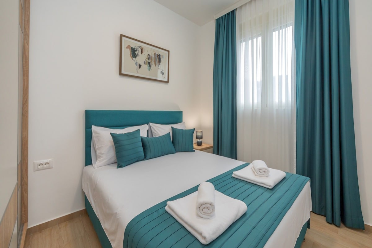 Habitación de hotel con cama, cortinas color turquesa, un cuadro en la pared y una mesita de noche con lámpara.