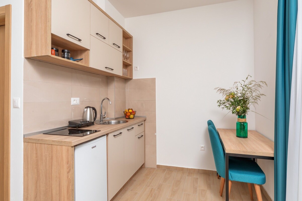 A compact kitchen with wooden cabinets, a sink, a kettle, and a bowl of fruit. There is a dining table with a chair and a vase of flowers.