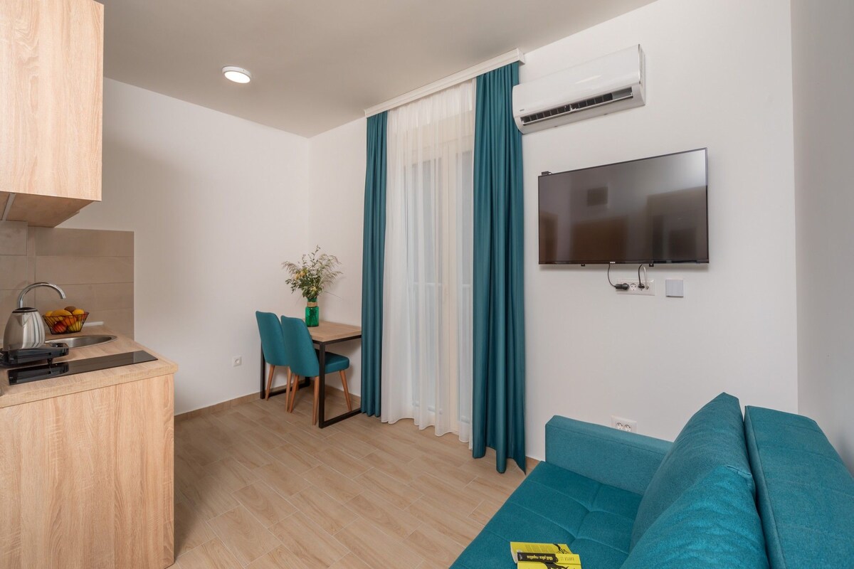 A modern living room with a teal sofa, a flat-screen TV mounted on the wall, and a dining area with a table and chairs.