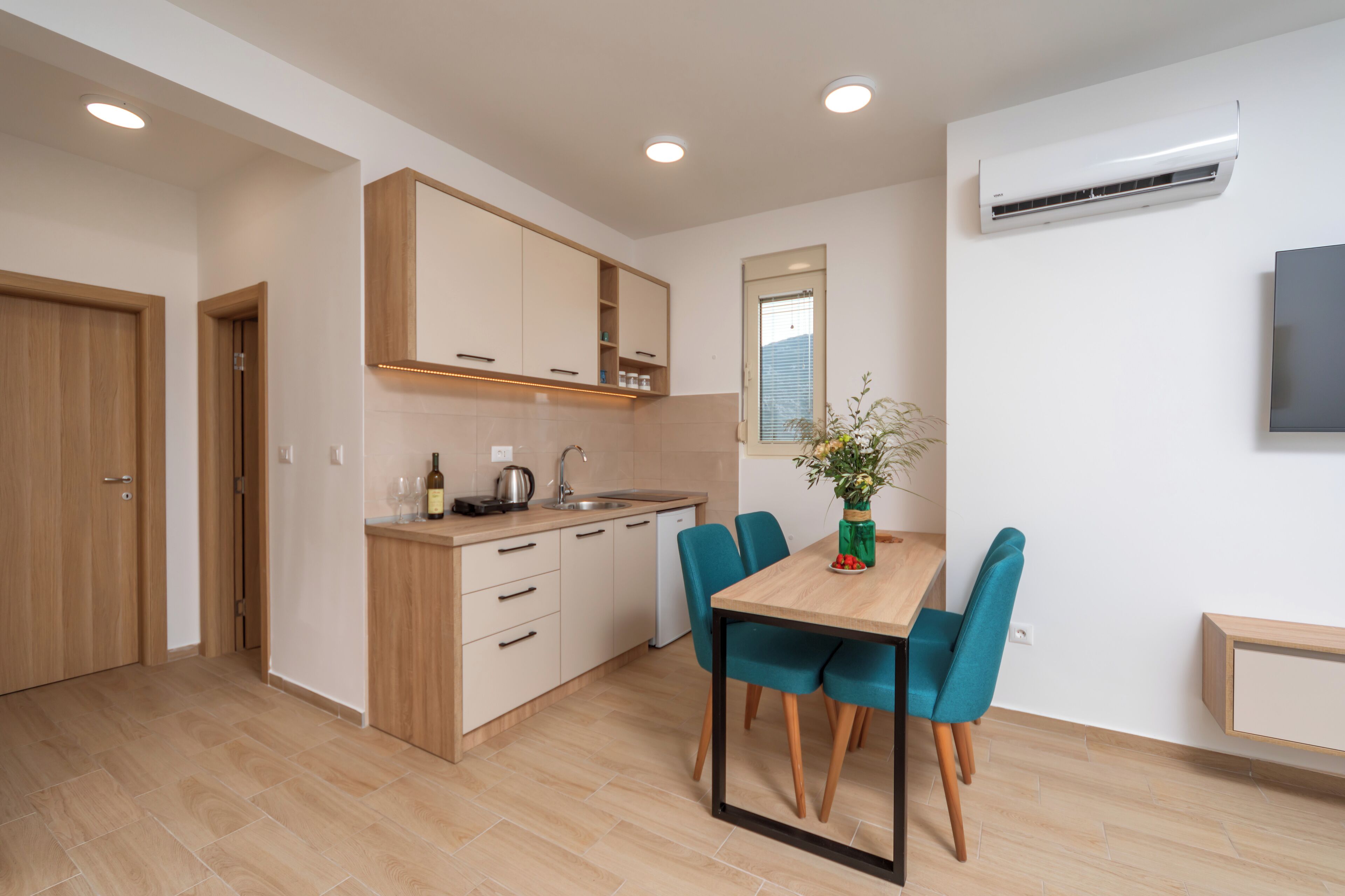 A modern kitchen with wooden cabinets, a built-in oven, and a dining area with a wooden table and teal chairs.