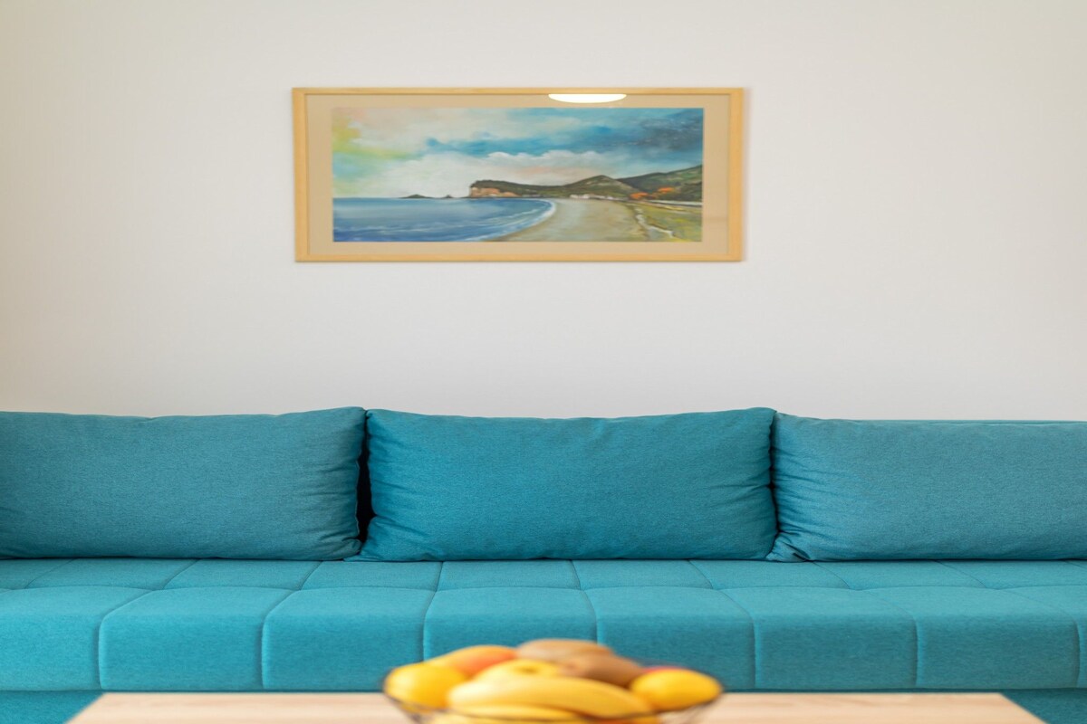 Un sofá azul aguamarina con un cuadro de una playa sobre él. Delante del sofá, hay una mesa de madera con un cuenco de fruta.