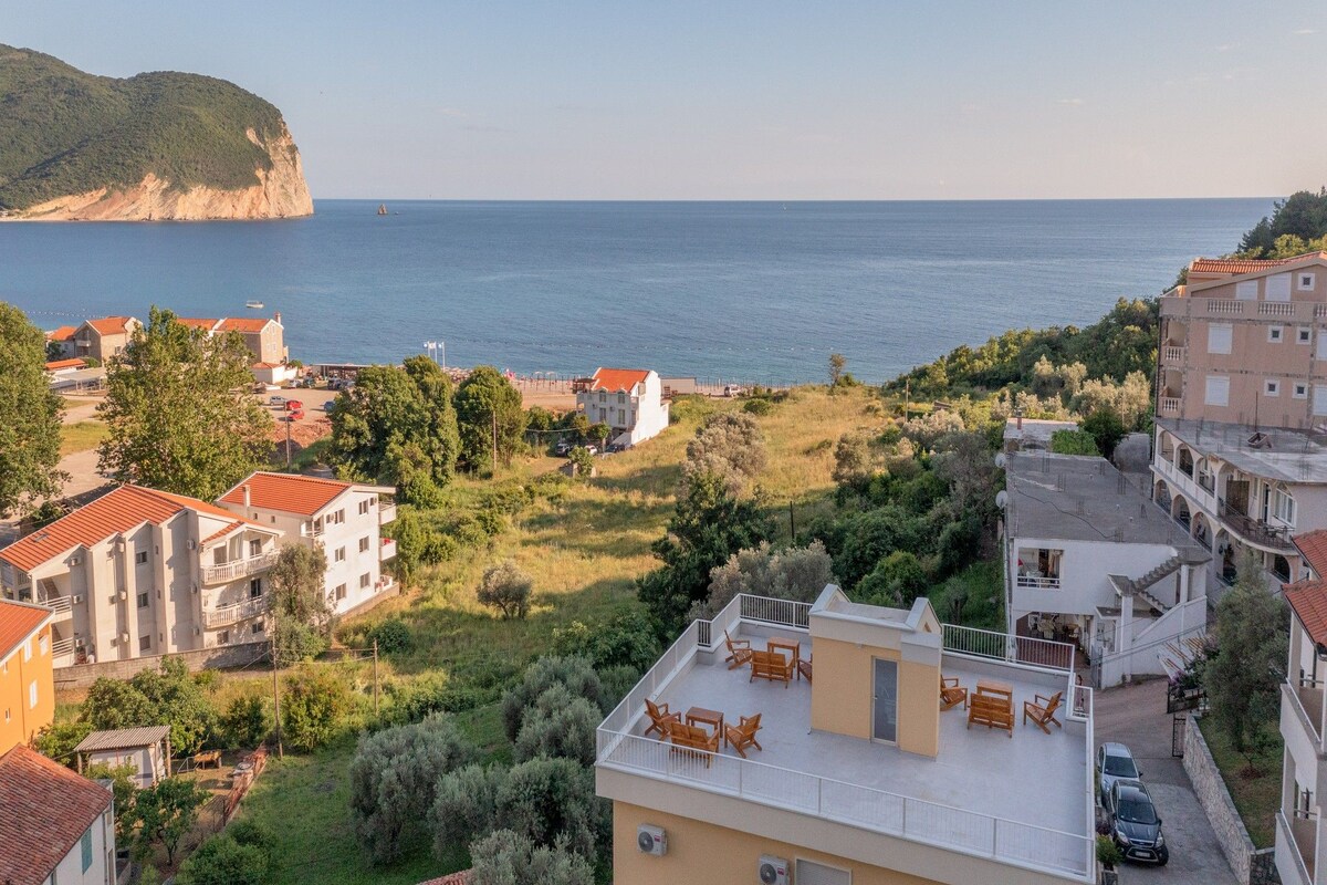 A coastal residential area with buildings, a view of the sea, and a hillside.