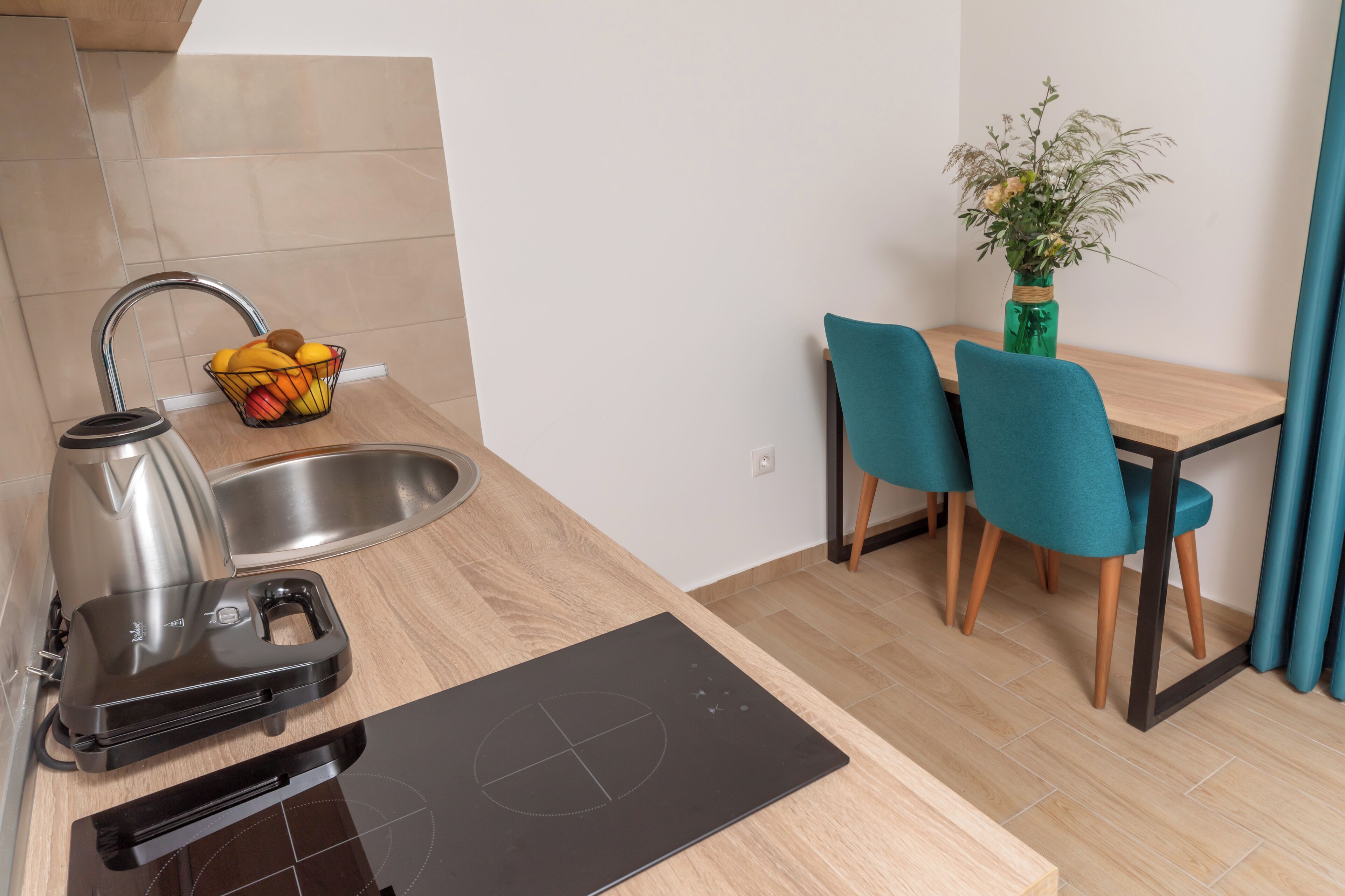 Une cuisine compacte équipée d’un évier, d’une bouilloire et d’une plaque de cuisson à induction. Un panier de fruits est posé sur le comptoir, et il y a une petite table avec des chaises, ainsi qu’un vase rempli de fleurs.