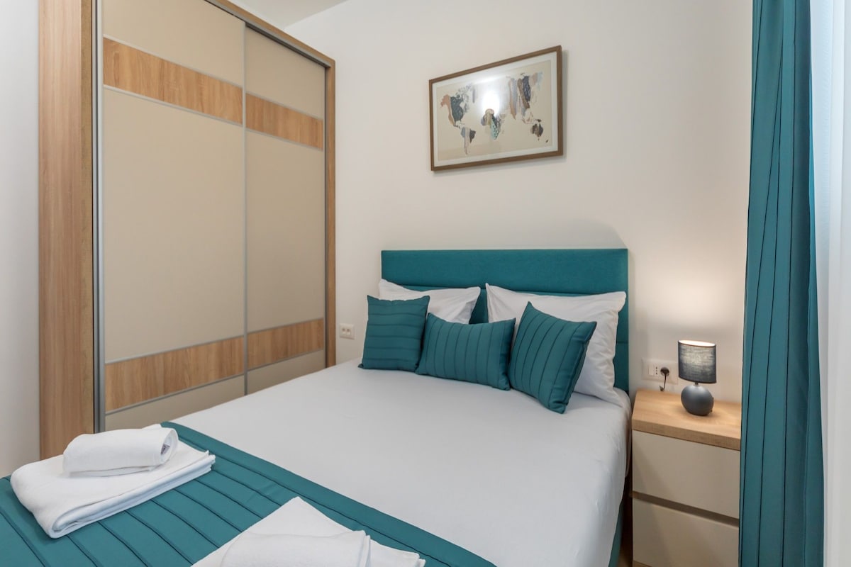 A neatly made bed with teal bedding and pillows, a wooden wardrobe, a bedside table with a lamp, and a world map artwork on the wall.