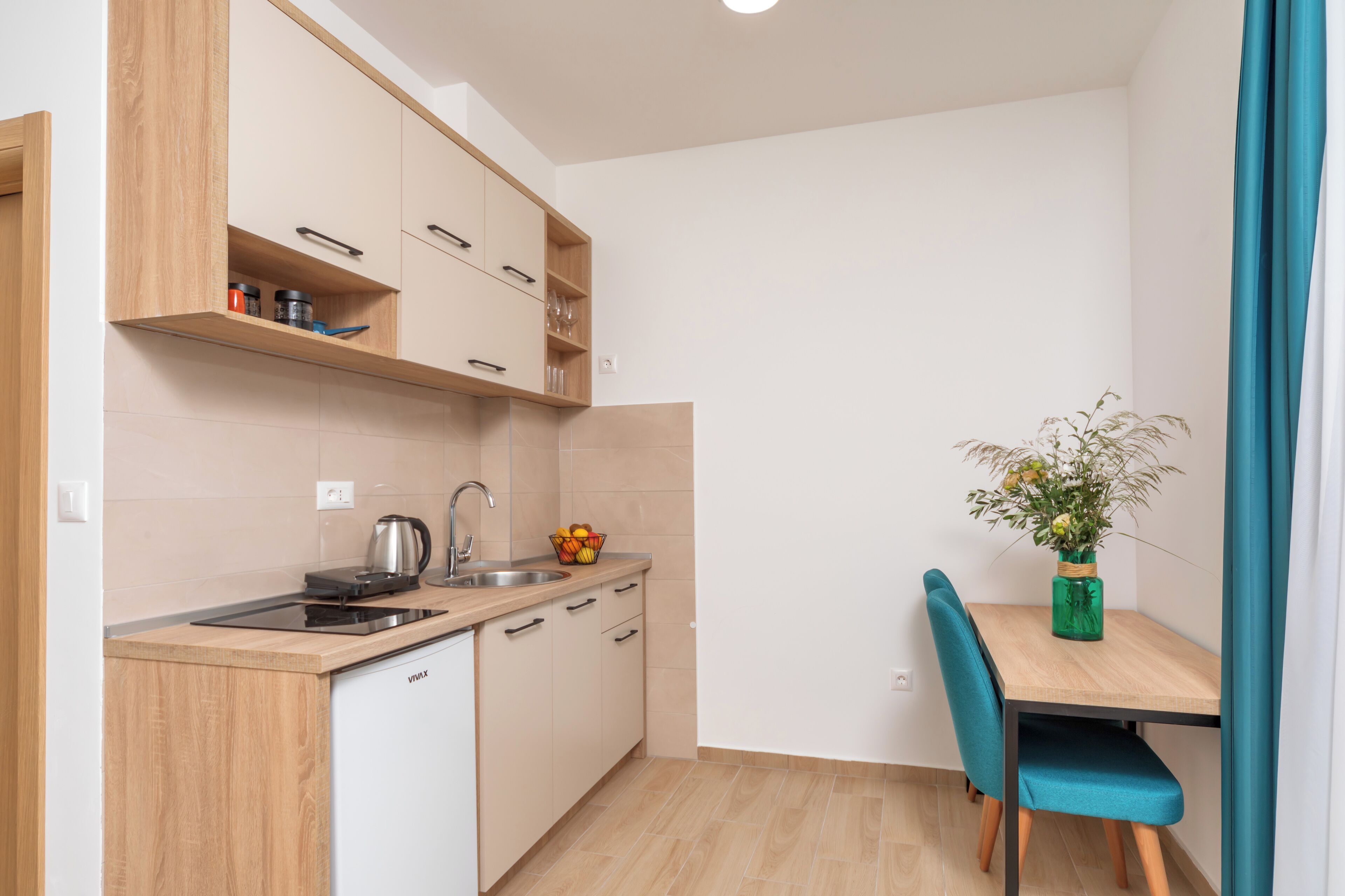 A compact kitchen with a dishwasher, a sink, and a countertop with a kettle. There is a small table with a vase of flowers and a chair.